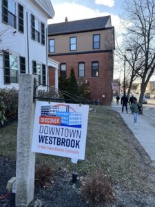 A sign says "Discover Downtown Westbrook, a Main Street America community." Diverse people are walking on sidewalks in a New England historic downtown business district. 